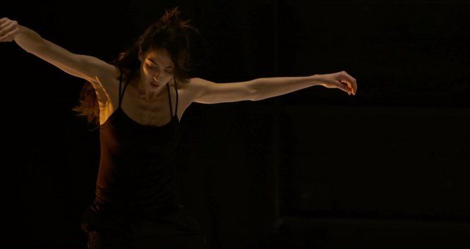 jeune femme danse debout les bras en croix devant un fond noir